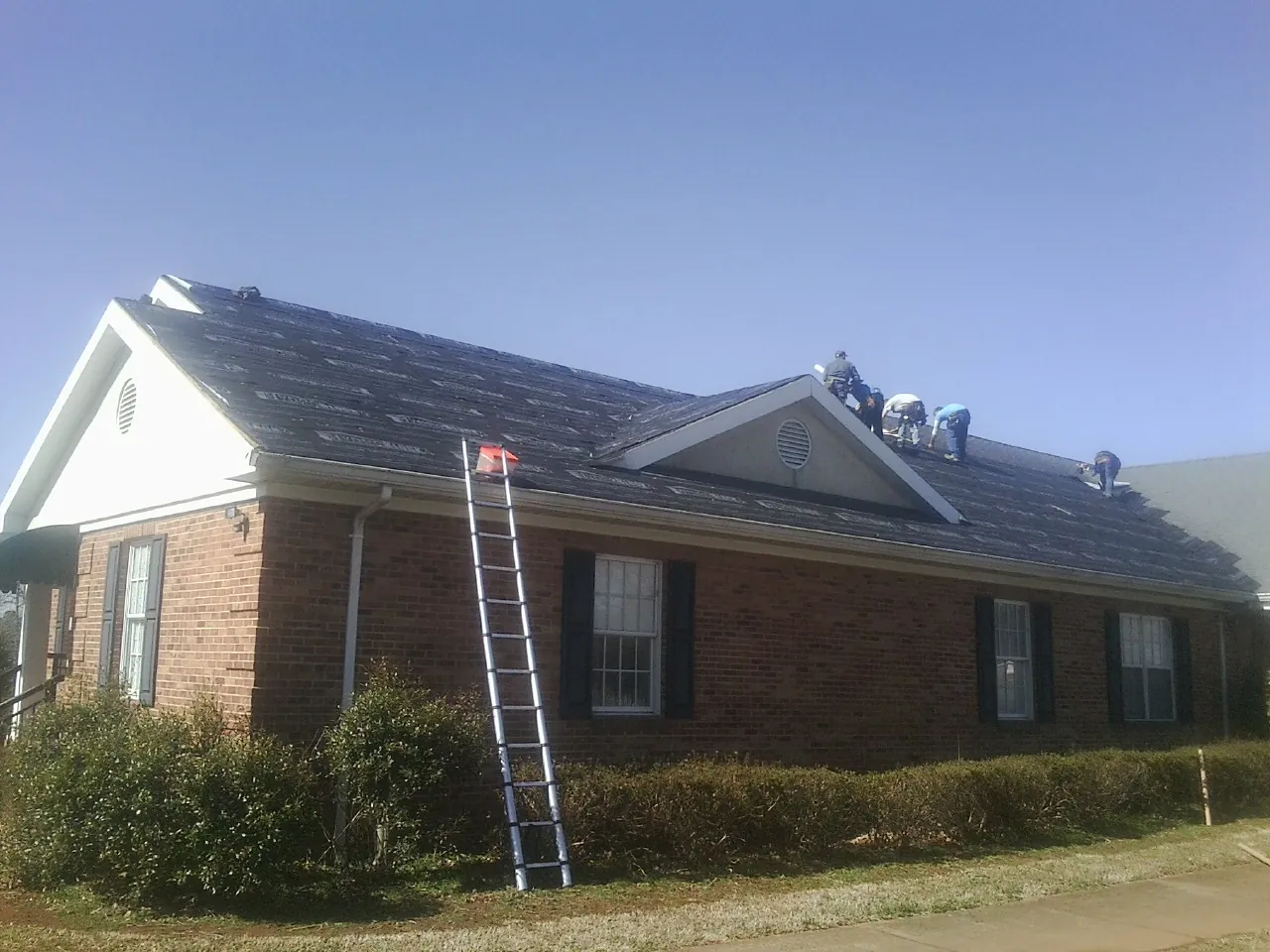 Crew tearing shingles off a brick ranch roof with a ladder set — Metal Roof Repair work by our crew in Alvada