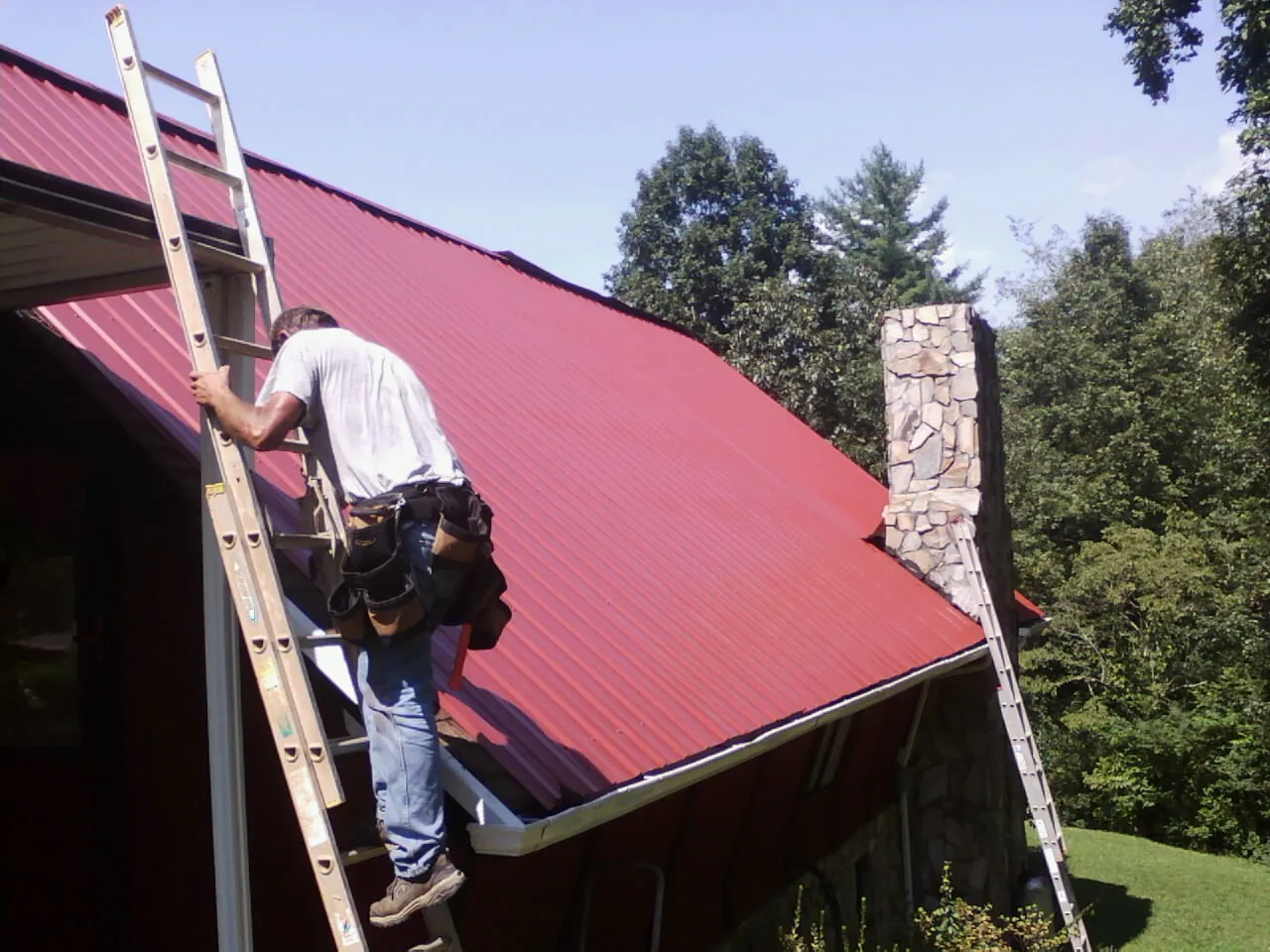 Roofer on a ladder beside a red metal roof and stone chimney — Soffit & Fascia Repair by Roofing Brothers in Alvada